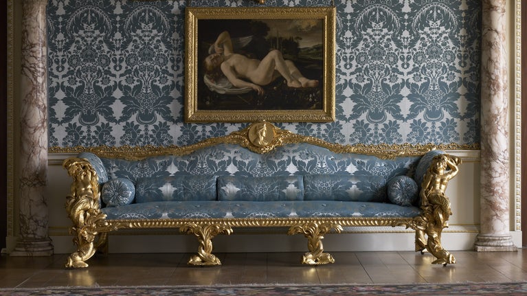 Large blue jacquard and gold chair with matching wallpaper behind and a nude painting above